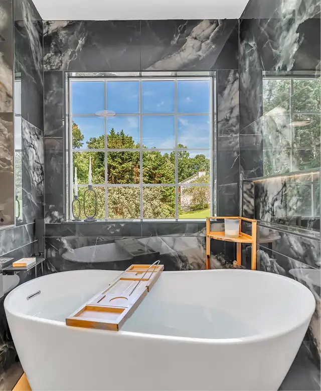 Freestanding soaking tub under a picture window framed by dark marble-look wall tile
